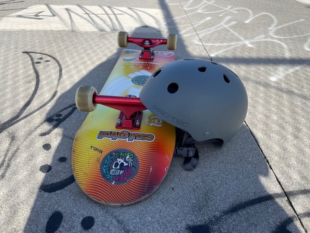 A photo of a Pro-Tec skateboard helmet leaning against a skateboard at a skatepark. Random graffiti is visible. 