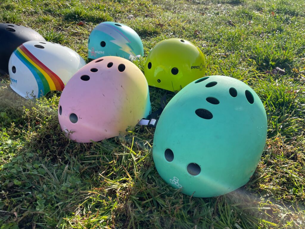 A collection of colorful skateboard helmets sitting on the grass outside. They are different colors and different brands. 