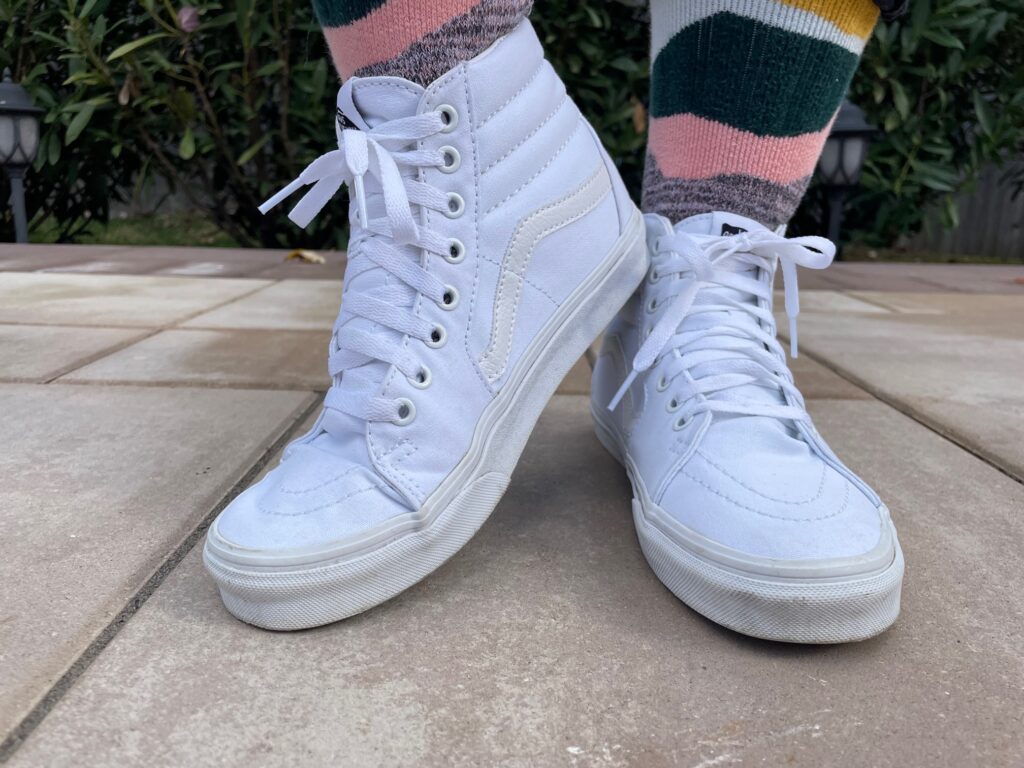 Two feet wearing Vans white high-top skateboard sneakers with the laces tied. The socks are very colorful. 