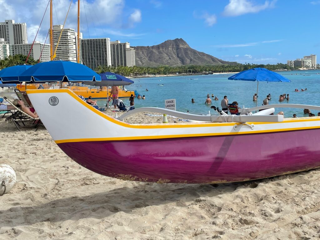 A red and white outrigger canoe on the beach and Diamond Head beyond. 