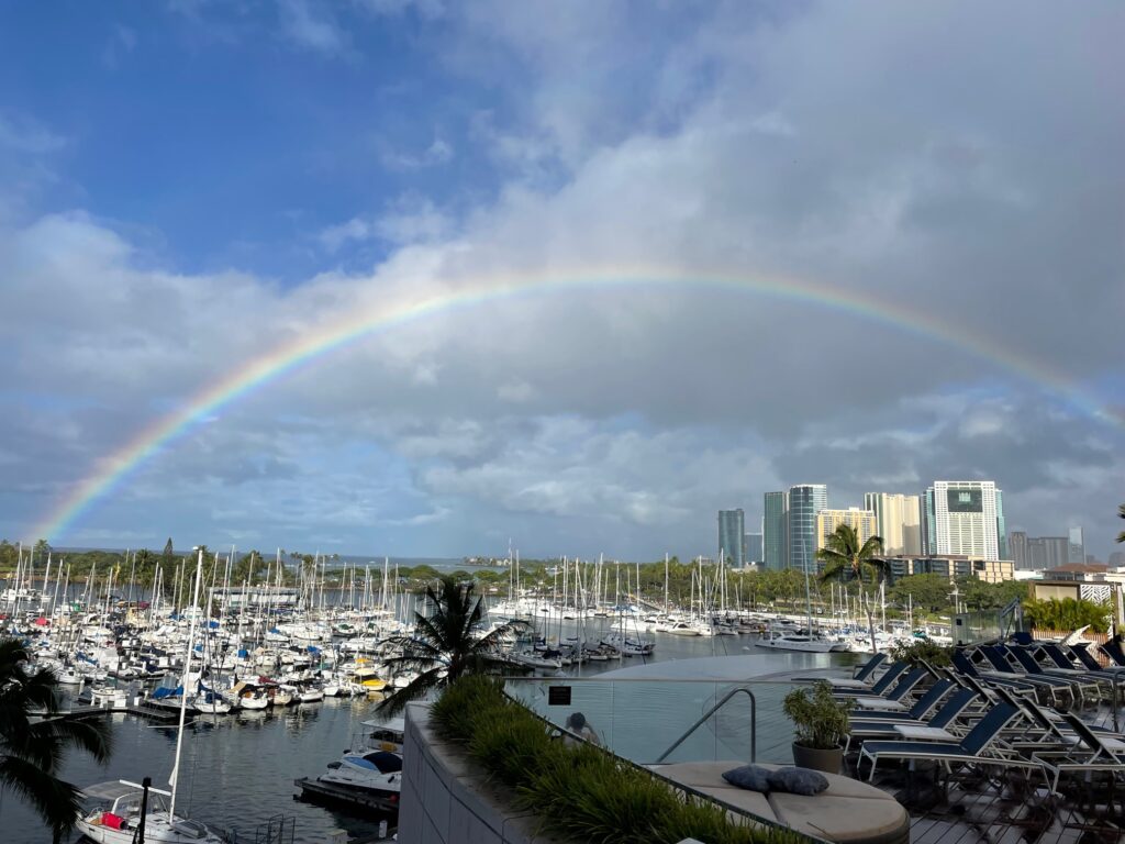 Rainbow over the marina in Waikiki, Hawaii. Many boats are in the marina and you can see high-rise buildings at a distance. 