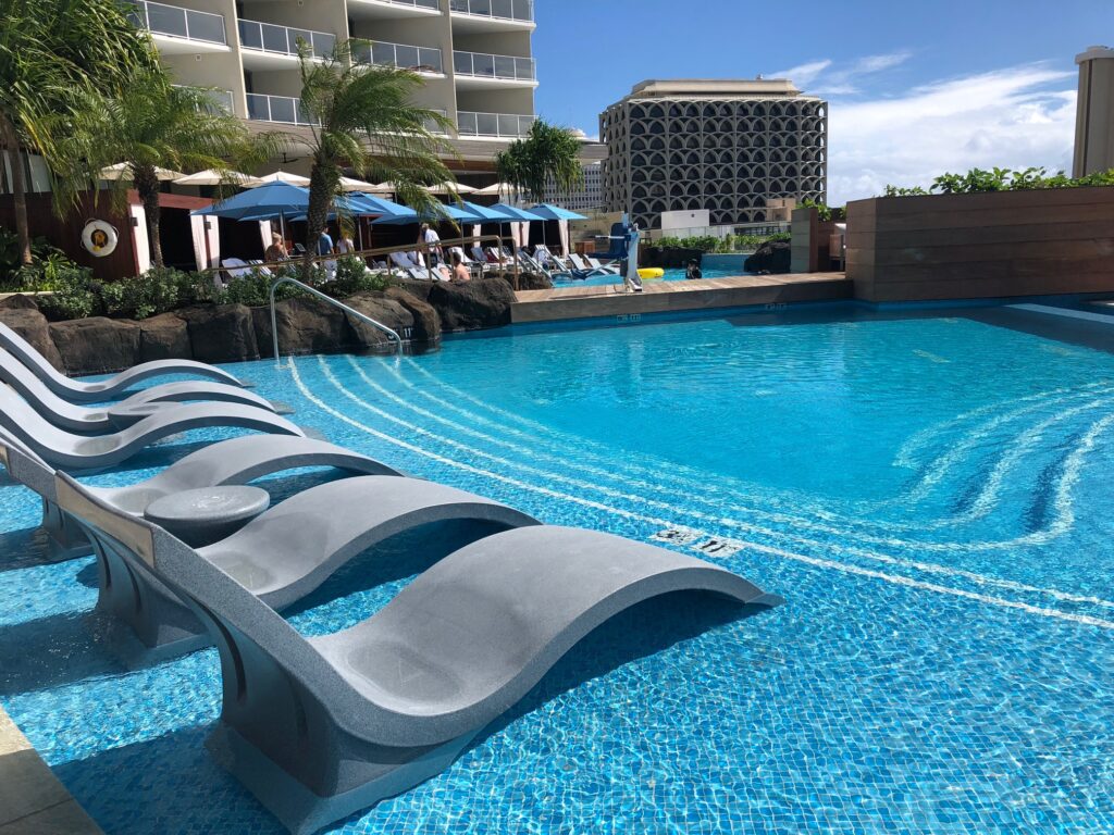 Ritz Carlton Waikiki pool with lounge chairs in the shallow water and buildings beyond. 