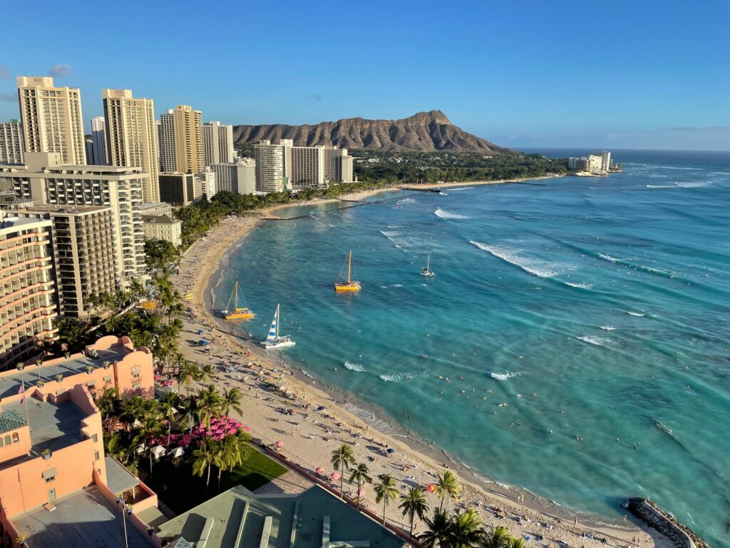 View of Waikiki Beach and Diamond Head from Sheraton Waikiki. The sky is blue and many high-rise hotels line the shore.