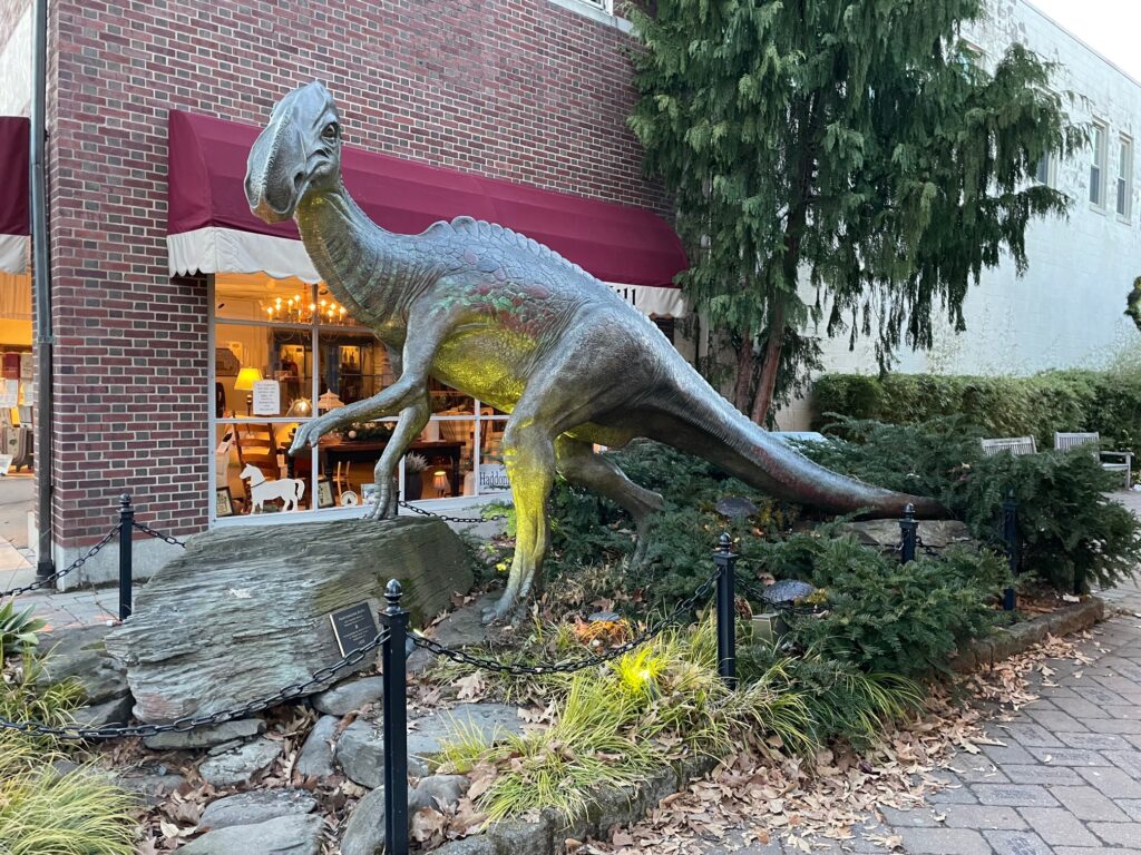 The bronze dinosaur statue located in the center of Haddonfield, NJ with a shop and trees behind it. 