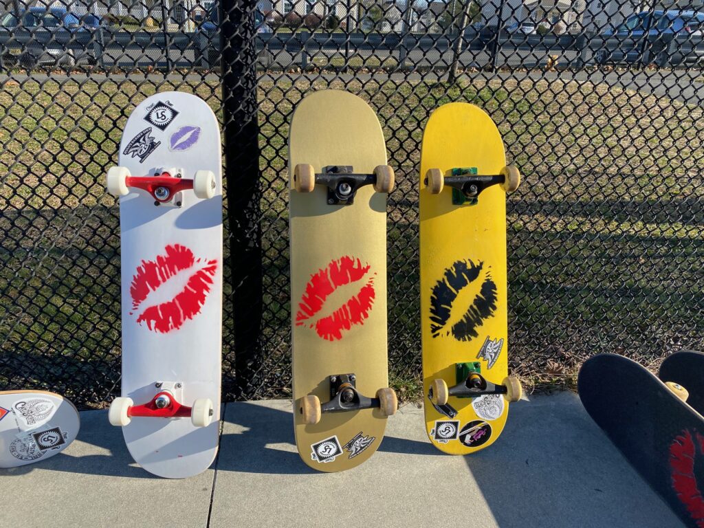 Three skateboards made by the Lipstick skate company lined up against a chain link fence. The boards have lipstick imprints on them. 