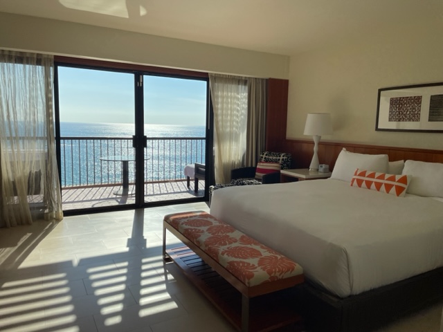 View of a guest room at the Mauna Kea resort with a king bed and an ocean view beyond. 